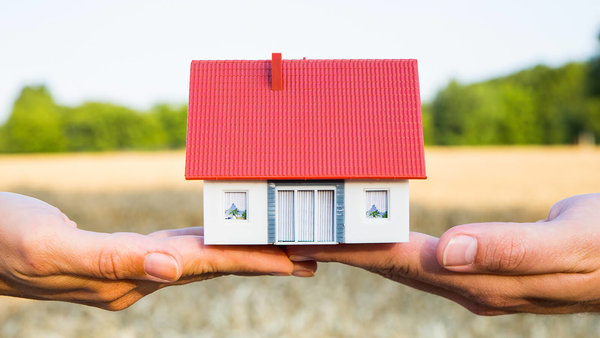 Zwei Hände halten über einem Kornfeld ein Modellhaus.